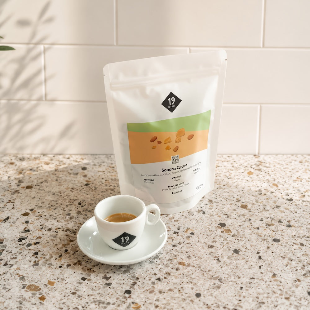 19grams Sonora Caturra Espresso Mood-Shot – Kaffeebeutel hinter einer Espressotasse mit 19grams Logo auf einer Terrazzo-Arbeitsplatte