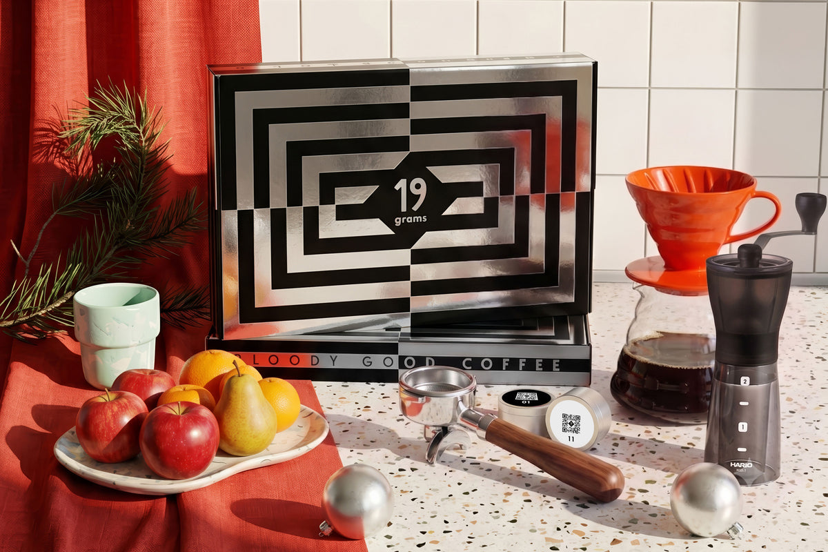 19grams Adventskalender 2026 – Moodbild der schwarz-silbernen Kalenderbox „Bloody Good Coffee“ mit Siebträger, Kaffeedosen, orangem Handfilter, Handmühle und Obstteller vor rotem Vorhang