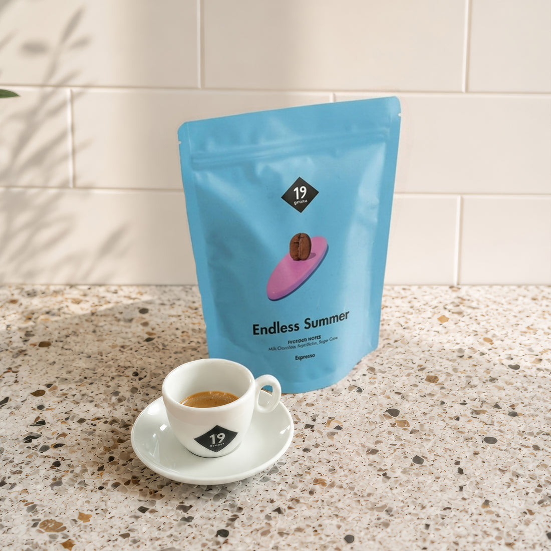 19grams Endless Summer Espresso – hellblauer Kaffeebeutel mit Kaffeebohne auf pinkem Surfbrett neben einer Espressotasse auf einer Terrazzo-Arbeitsplatte