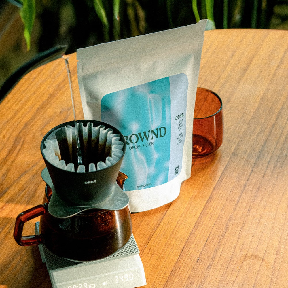 19grams GROWND Dusk Filter Decaf – Moodbild mit Kaffeebeutel auf Holztisch neben Handfilter, der auf einer Waage in eine Glaskaraffe aufbrüht, und braunem Trinkglas