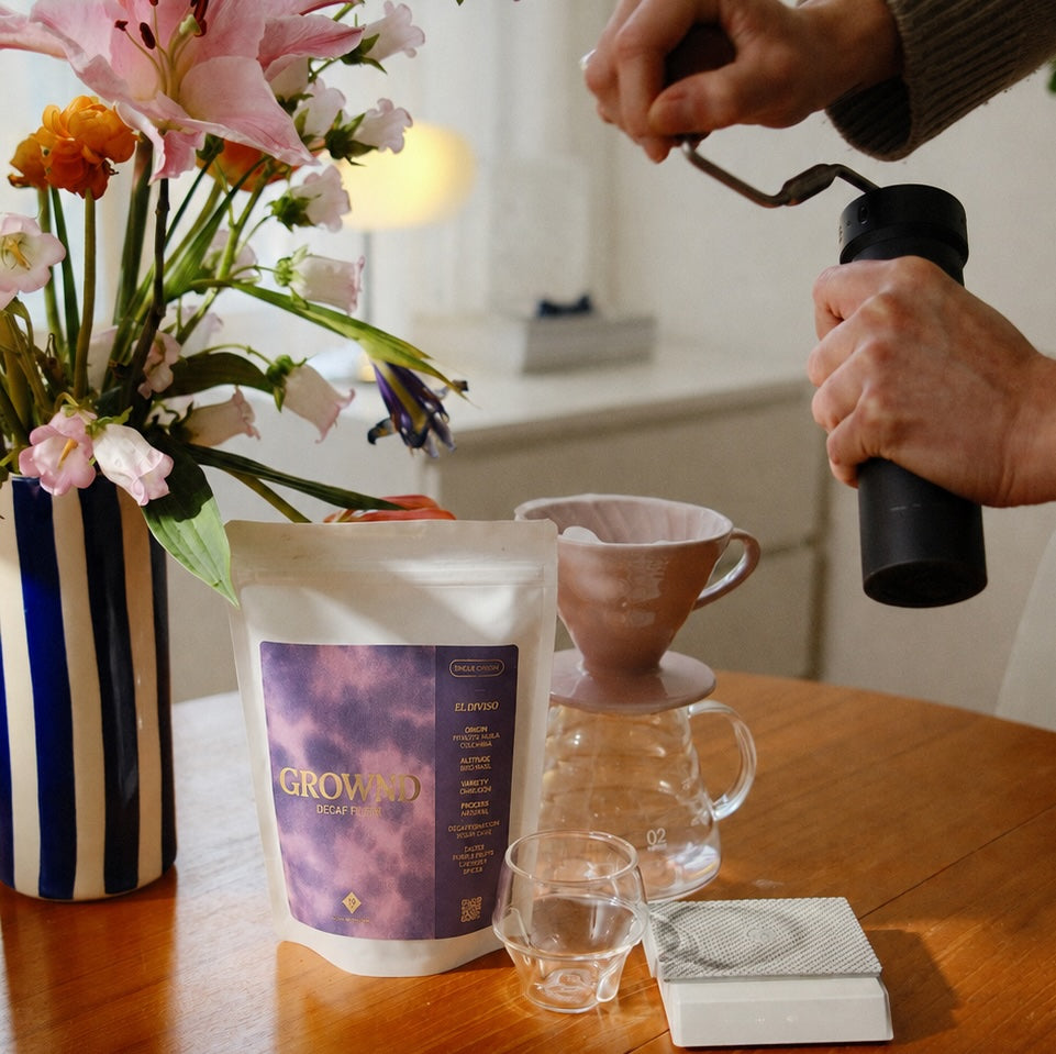 19grams GROWND El Diviso Decaf Filter – Moodbild mit Kaffeebeutel auf Holztisch, rosa Keramik-Handfilter auf Glaskaraffe, Handmühle und gestreifter Blumenvase
