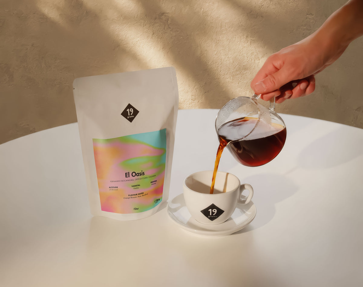 19grams El Oasis Filter – Kaffee wird aus einer Glaskaraffe in eine Tasse gegossen, daneben der Kaffeebeutel mit pastellfarbenem Verlaufslabel
