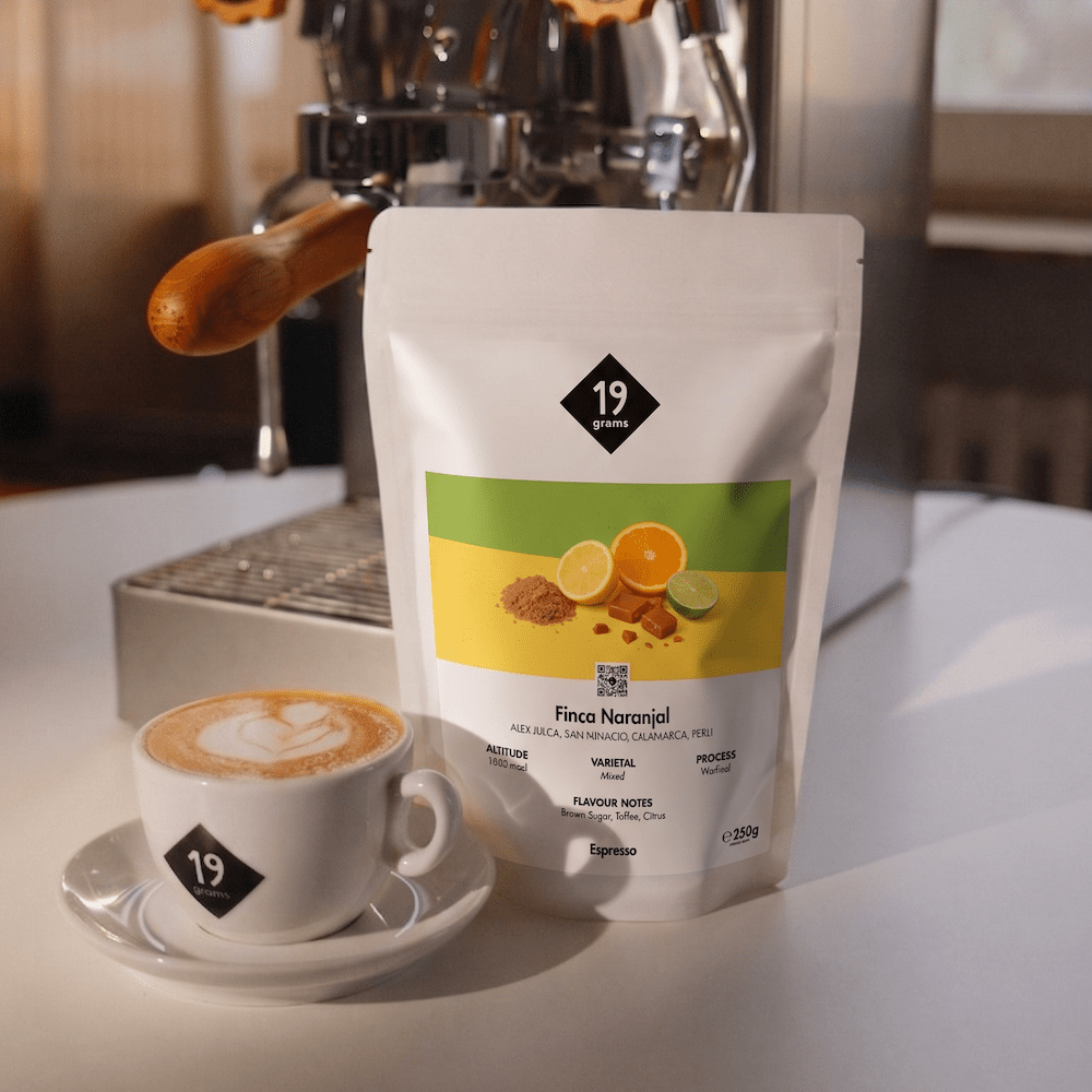 19grams Finca Naranjal Espresso Kaffeebeutel vor Espressomaschine mit Espressotasse und Latte-Art – Specialty Coffee Peru Moodshot
