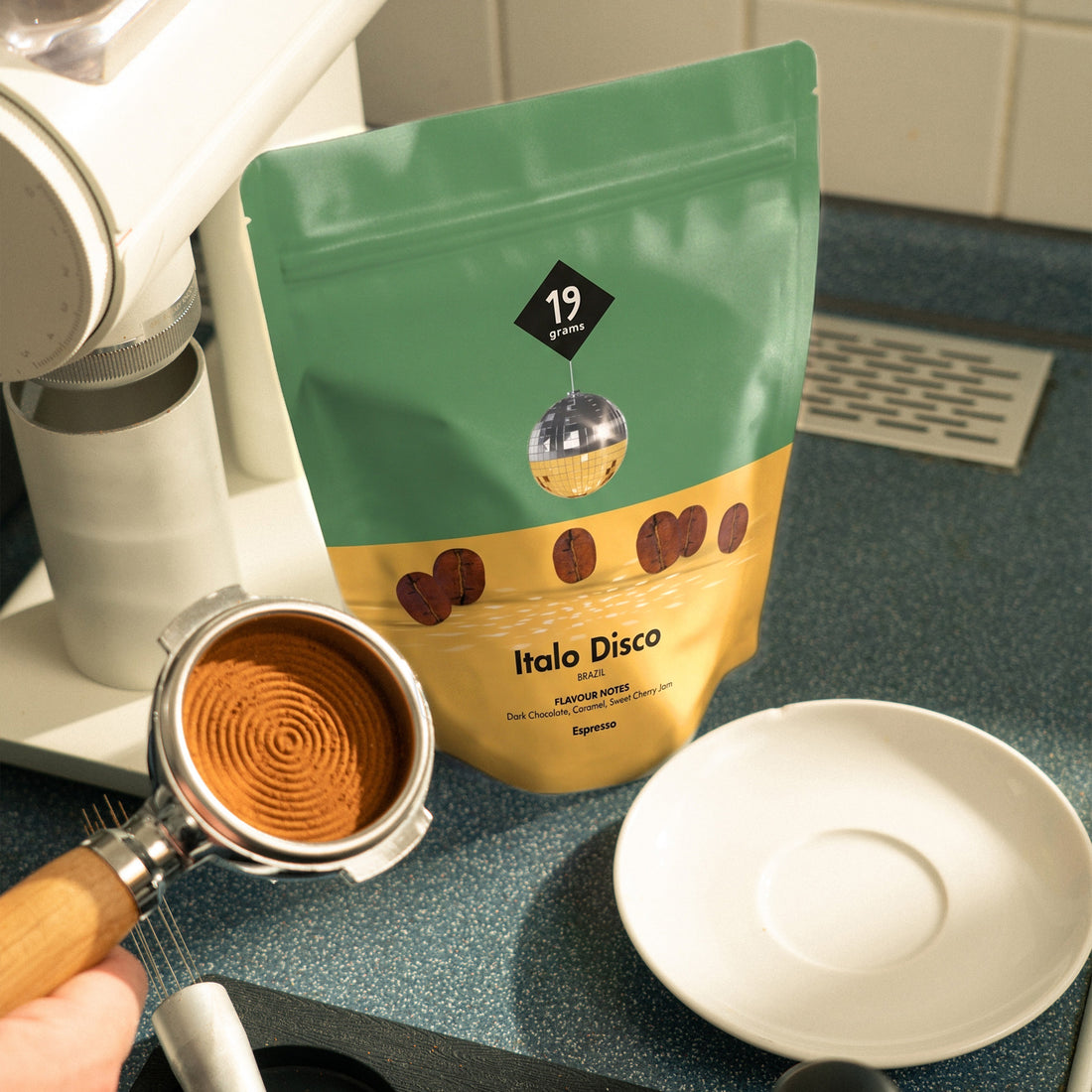 19grams Italo Disco Espresso – grün-goldener Kaffeebeutel mit Discokugel neben einem gefüllten Siebträger und einer Espressotasse auf einer Küchenarbeitsplatte