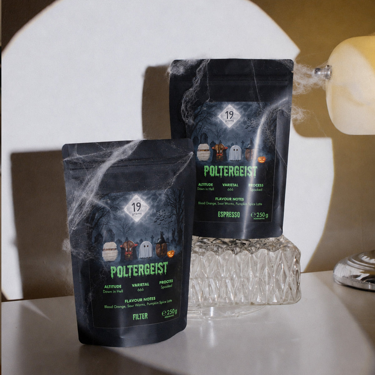 Zwei 19grams Poltergeist Kaffeebeutel, Espresso und Filter, mit Kunstspinnweben dekoriert auf einer Anrichte neben einer Glasschale und einer Lampe
