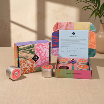 19grams The Fermentation Project Kaffee Kit Espresso – Moodbild mit geschlossener marmorierter Geschenkbox, geöffneter Kaffeebox mit vier Dosen und Löffel sowie Pflanze auf beigem Untergrund