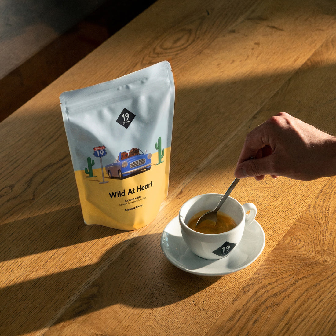 19grams Wild At Heart Espresso – hellblau-gelber Kaffeebeutel mit blauem Cabrio an der Route 19 zwischen Kakteen, daneben eine Espressotasse auf einem Holztisch